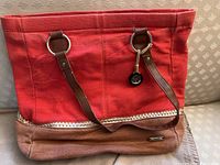 Photo of a red and tan canvas tote bag with brown straps and metal rings, lying on a patterned beige couch.