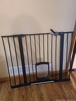 Black metal baby and pet safety gate with lockable clear plastic pet door and attached extender pieces, shown placed against a beige wall and hardwood floor.