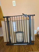 Black metal baby and pet gate with lockable pet door placed upright against the wall.