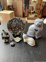 Overview image of all cat-themed decorative items on table including plate, large cat figurine, white cat figurine, cat ashtray, and three small cat figurines