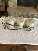 Three cream ceramic kitchen canisters with lids displayed on a decorative black metal scrollwork stand. Each lid is labeled for Sugar, Coffee, or Tea.