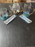 Full view of decorative tea set including teapot, two mugs, spoons, cloth napkins, and coasters arranged on black surface