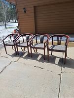 Four wooden side chairs with fabric cushions pictured outdoors on a concrete patio near a house.