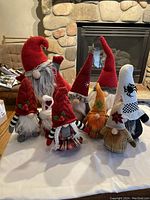 Nine gnome figurines arranged before a stone fireplace, showing variety in hat colors including red, white, and orange, and various holiday designs.