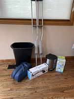 Full view of all items in the lot including adult crutches, Pure Wave massager box, Easy Feet foot cleaner box, black plastic trash can and small stainless steel step trash can all placed on wooden floor in front of window