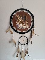 Front view of the dream catcher showing the deer scene artwork inside the black hoop with feathers and beads hanging down.