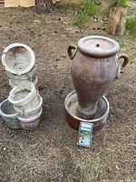 Two outdoor water fountains placed on ground outdoors, one is a tall urn-shaped fountain with handles and a wide lip, approximately 32 inches tall, brown finish. The other is a smaller stacked bowl fountain with grey textured finish. Fountain pump packaging visible in front.