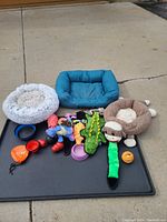 Photo of two pet beds, one round grey plush and one square teal, along with a brown sloth-shaped pet bed, various dog toys including rubber balls, stuffed animals and chew toys, all arranged on a large black kennel tray.