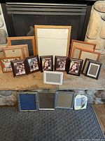 Photo showing twenty picture frames in mixed materials including wood, metal, and ceramic arranged on a stone hearth and carpeted floor.