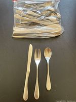 Photo showing 12 place settings of silverware including knives, forks, and spoons. Silverware is arranged with some items loose and some in a plastic bag.