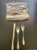 Photo showing three types of silverware pieces (knife, fork, spoon) arranged in front of a sealed plastic bag containing multiple similar pieces.