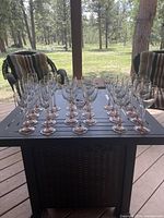 Photo showing all 23 rose stem wine glasses arranged in rows on a dark outdoor table with two cushioned chairs in the background.