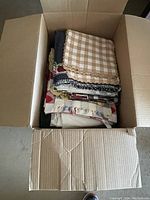 Open cardboard box containing a stack of folded assorted table linens including placemats and tablecloths in various colors and patterns