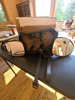 Pair of towing mirrors with black plastic housing and rubber straps lying on a table with a cardboard box behind them. Sunlight through windows reflects in the mirror surfaces.