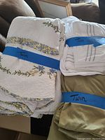Three folded twin sized bedding items: white quilt with floral embroidery and blue floral border, light comforter with striped pattern, and solid olive green sheet or cover.