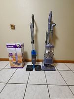 Photo shows three cleaning appliances: a boxed new Shark steam mop on the left, a blue Bissell steam mop with twin square mop pads in the middle, and a Shark Navigator upright vacuum cleaner on the right.
