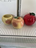 Front view of three ceramic cookie jars: one large red apple shaped with lid, one round yellow-to-pink gradient with lid, and one round yellow-to-pink gradient without lid.