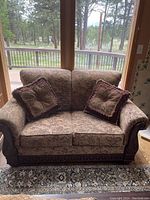 Vintage upholstered love seat with beige and brown patterned fabric, carved dark wooden trim, and two matching throw pillows with fringe trim shown in front of windows.