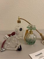 Three antique glass perfume bottles displayed on a flat surface, showing shapes and details including atomizer bulb and floral spray pump details.
