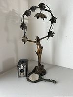 Full view of Art Deco lady lamp with extended floral arms showing non-operational lamp shaded fixture, cast metal hand mirror angled in front, and Kodak Brownie camera to the side on a white surface.