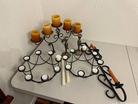 Large tiered black metal candle holder with five yellow candles, two black metal wall mount candle holders, two black metal eight-candle holders, and two silver candlestick holders with some candles.