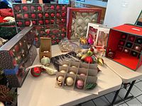 Large collection of boxed and loose Christmas ornaments on a table including round balls, bird-shaped ornaments, and glass icicles, with an ornament organizer.