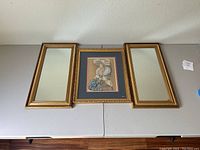 Photo shows the mixed media collage of a woman wearing a hat framed in gold with dark blue mat, flanked by two gold framed mirrors on each side, positioned against a neutral wall.