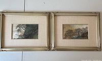 Two framed embroidered silk landscape artworks side by side on a flat surface with light beige matting and gold-tone frames.