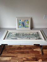 Two framed oil paintings on a table against a wall, one large horizontal cityscape and one smaller square cafe scene.