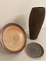 Plate with brown and white figurative design leaning against wall, geometric patterned plate, and tall dark brown polyresin vase with diamond design placed on floor