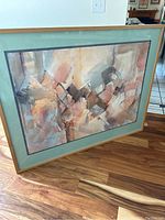 Photo of the framed abstract painting showing geometric and freeform shapes in muted tones of beige, peach, brown, gray, and white. Signature Tiret 89 visible in lower left. Mounted with pale green mat and under glass in frame.