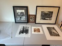 Photo showing six works of art displayed on table and leaning against wall, includes framed and unframed prints ranging from black and white landscape and architectural scenes to smaller etching.