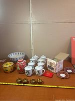 Full lot overview showing large white bowl, yellow lidded bowl, red covered jar, six white teapots, four brass cups, decorative plates.