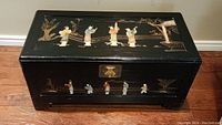Rectangular black lacquered wooden chest with hinged lid, metal latch, mother-of-pearl inlay figures and landscape motifs.