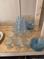 Blue tinted Depression Glass lot arranged on wood surface including pitcher, stemmed glasses, coupe glasses, and small plates.