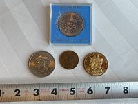 Four coins displayed on fabric with measuring tape for scale, showing various sizes and designs relevant to British Royalty.