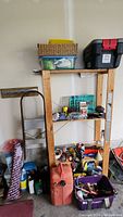 Wide view of wooden shelf filled with garage and workshop items such as tools, lubricant bottles, gas can, toolbox, and small plastic container with organized parts.