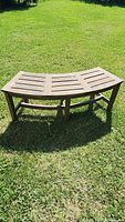 Curved teak bench on grass showing full length and slatted seat design from a side angle.