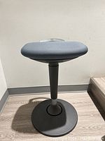 Front view of Starling Stool showing circular black base, central column, and grey upholstered seat.