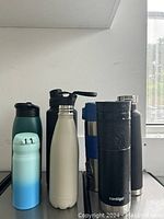 Photo showing seven metal water bottles in various colors and lid styles arranged in a row on a black surface against a plain white wall.