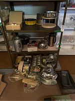 Shelving unit with three shelves holding various kitchen items including coffee percolator, tea kettle, metal bakeware pans, Wilton cake molds, boxed kitchen utensils, various packaged kitchen gadgets, and a wooden cheese board with tile inlay set on floor