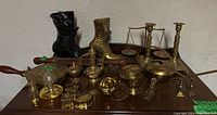 Wide view of lot showing black and brass boot figurines, brass bed warmer, candlesticks, balance scale, and miniatures spread on wooden surface.