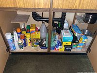 Overview of various cleaning supplies, food packaging products, pet supplies, and miscellaneous items stored under a kitchen sink on a wooden shelf with plumbing pipes visible.