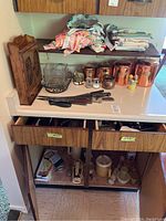 Wide view of cabinet countertop and drawers showing copper-tone canisters, cutlery, hand mixer, assorted linens, aprons and storage items.
