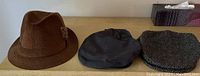 Three men's hats: brown corduroy fedora, dark grey cap, greyish herringbone flat cap, on wooden surface.