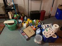 Wide view showing assortment of cleaning liquids in plastic bottles, paper goods including many toilet paper rolls, cleaning basket, mop, and cardboard box on floor.