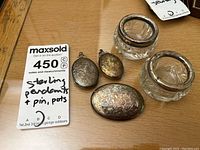 Photo showing three sterling silver lockets with engraved floral designs alongside two vintage crystal powder jars with silver tops on wooden surface.