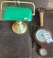 Photo showing green bankers lamp with brass base and Taylor weather station with mahogany frame containing barometer and thermometer.