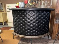 Front view showing curved black vinyl padded upholstery with tufted buttons, wood trim on top and bottom, and metal footrest along bottom.