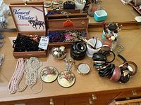 Photo showing multiple strands of costume bead necklaces, compact mirrors, wooden boxes with jewelry, and bangles on a wooden surface